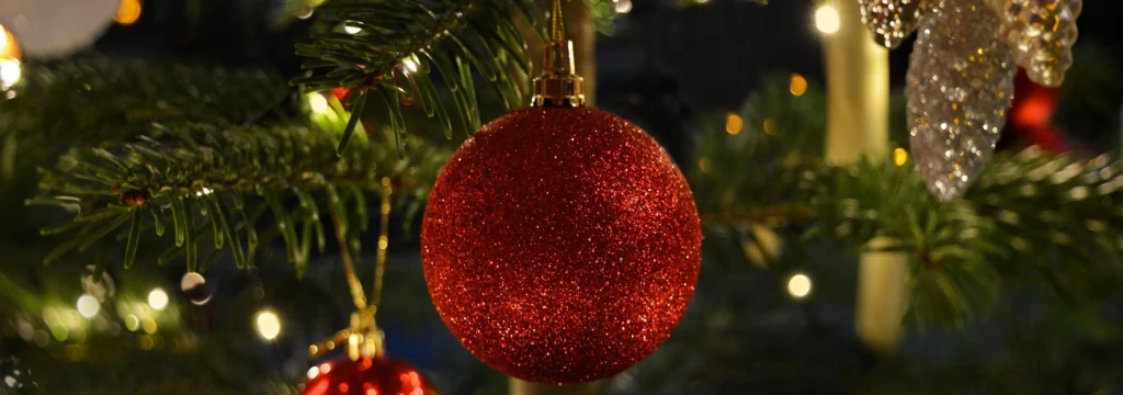 Rote Weihnachtskugel mit Glitzer hängt an einem Tannenzweig, umgeben von Lichtern und weiteren Weihnachtsdekorationen.