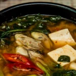 Koreanische Suppe mit Tofu, Muscheln, Gemüse und Gewürzen in einer schwarzen Schüssel, umgeben von frischen Kräutern.