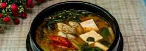 Koreanische Suppe mit Tofu, Muscheln, Gemüse und Gewürzen in einer schwarzen Schüssel, umgeben von frischen Kräutern.